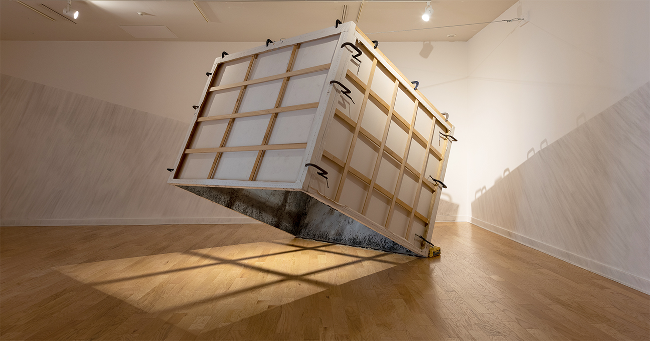 A shipping crate suspended in the air with a shadow being cast though it in a gallery.