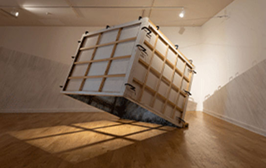 Tilted Heritage, 2015 A shipping crate suspended in the air with a shadow being cast though it in a gallery.