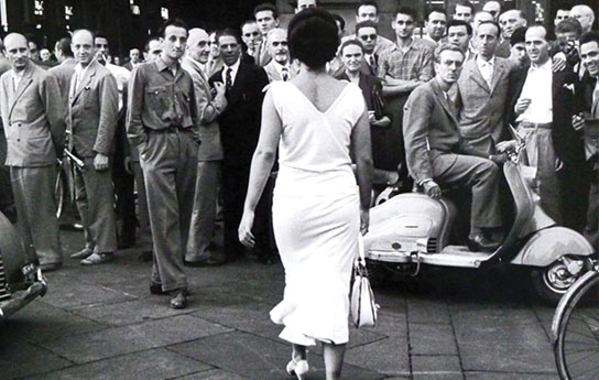 Thumbnail_Past (39) Women in a white dress walking into a crowd of men.