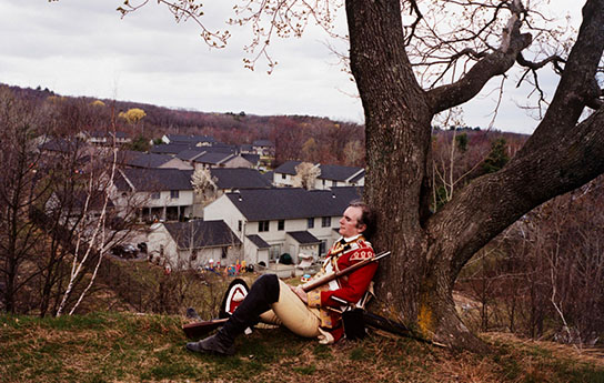 British Redcoat Re-Enactor, Battle Of Concord And Lexington, Lexington, Ma From Perfectible Worlds, 2002 A soldier sitting against the tree with a town in the distance